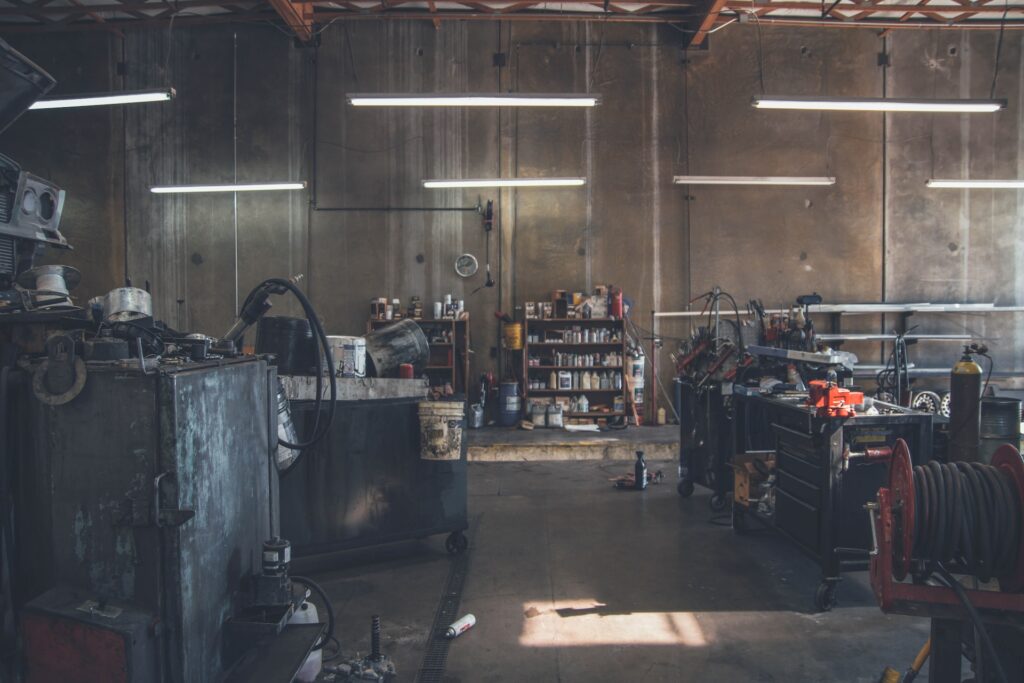 Different types of motor oil are organized on a brown wooden shelf placed near the dirty wall of an unkept auto repair shop