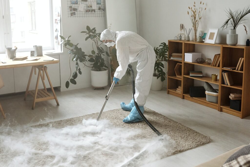 A person wearing white protective gear sanitizes the brown carpet near a wooden table and brown wooden shelf inside the white room