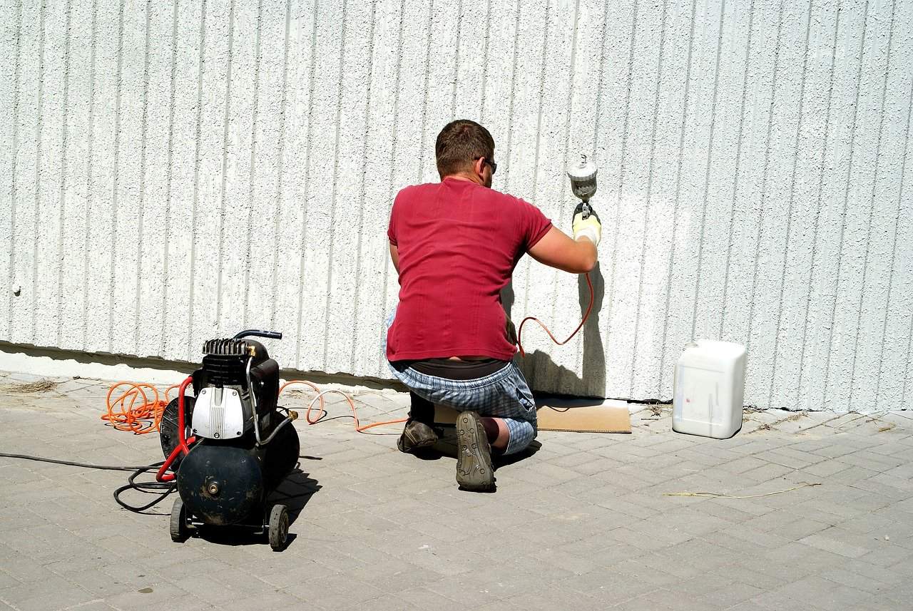 A man wearing a red shirt and blue checkered pants kneels on the pavement while spraying white paint on the wall