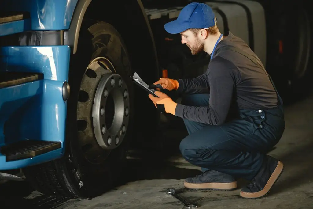 A man in a blue cap and blue working uniform is sitting beside a blue truck with a black wheel in a repair shop