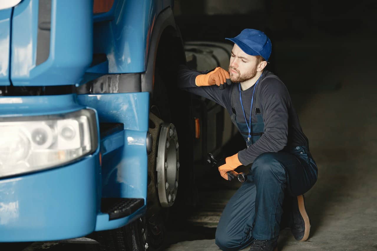 A worker in a blue cap and gray long sleeves shirt kneeling beside a blue truck in a repair shop