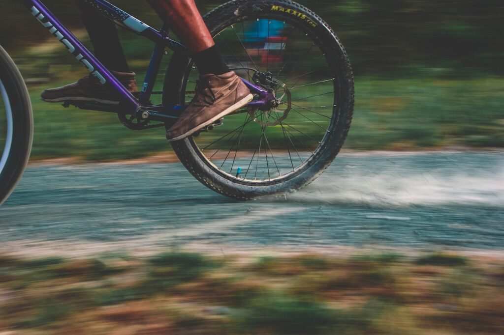 A person wearing black socks and brown shoes is riding a black and violet colored bike on a rough road