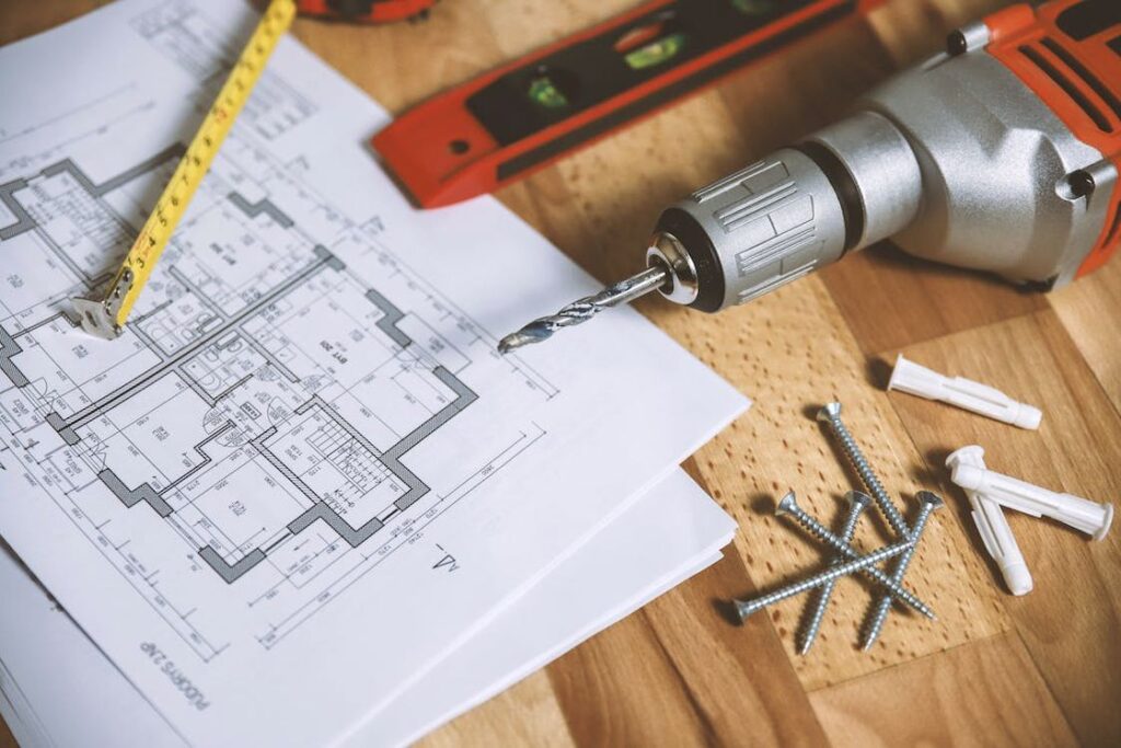 Drill with an attached drill bit is placed on a table alongside different woodworking tools and a floor plan