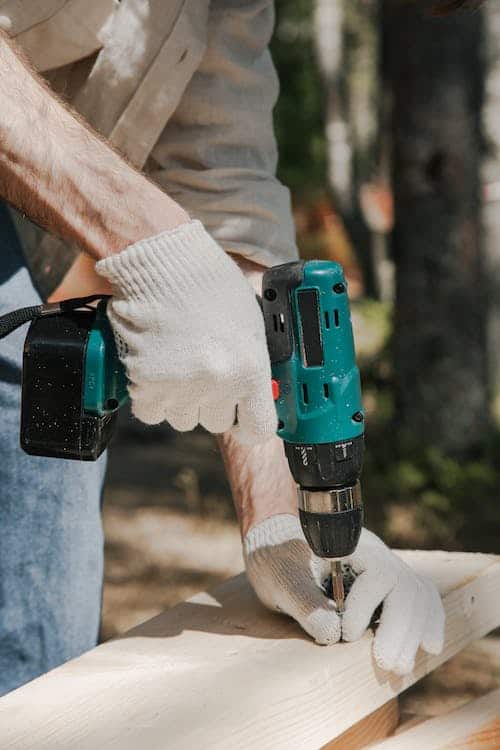 Man with gloved hands drilling a hole in a plank of wood