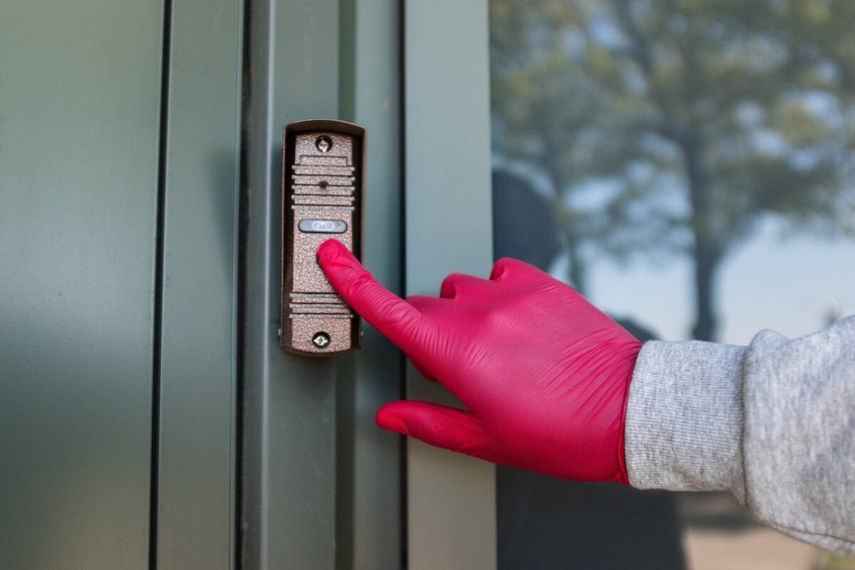 A person wearing gray long sleeves shirt and pink latex gloves is pressing the bronze-colored doorbell near the green door