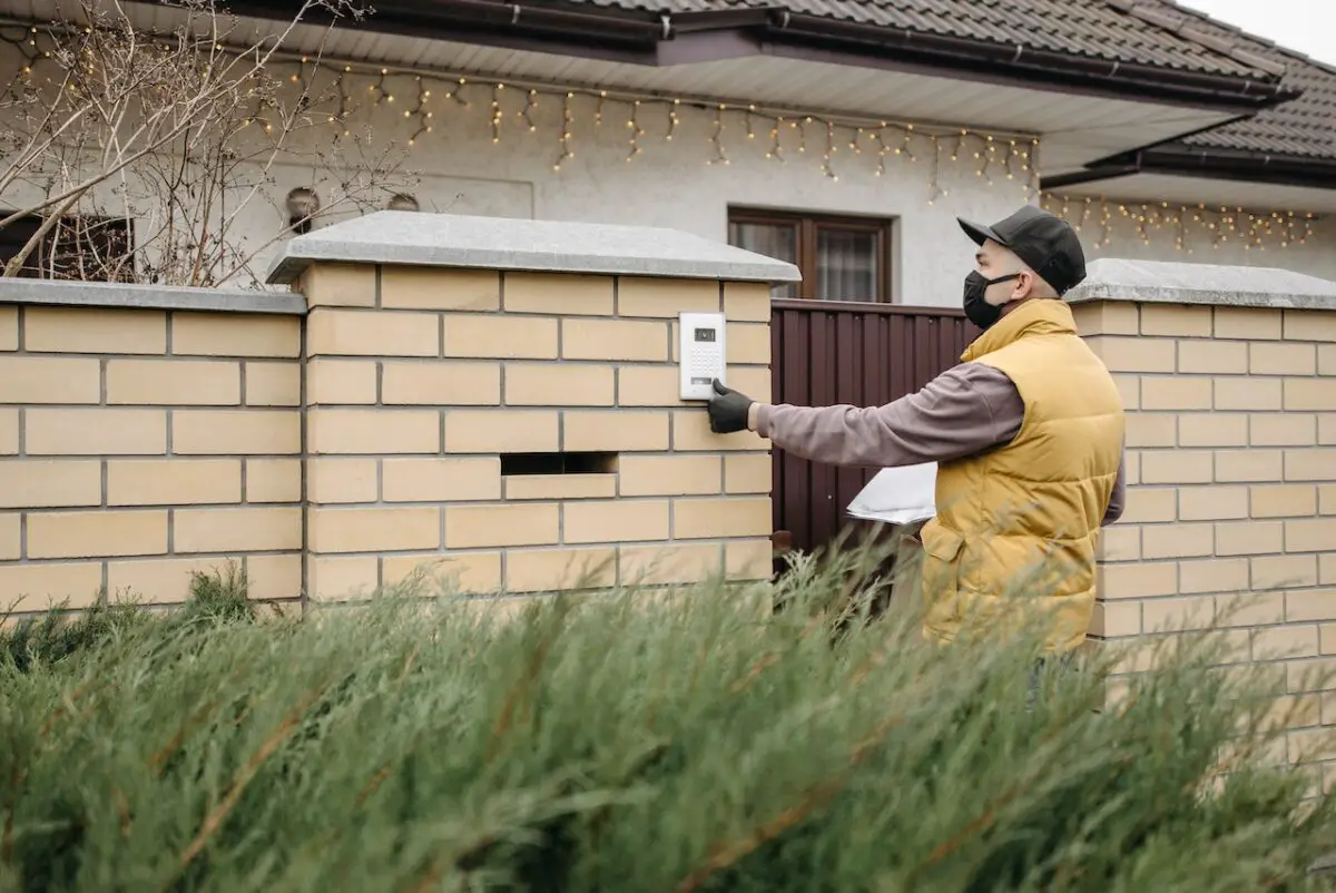 A man wearing a black cap, black mask, and yellow puff vest is pressing a white doorbell