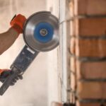 A man wearing a plain white shirt and black and orange gloves while using an angle grinder to cut fire bricks on a wall