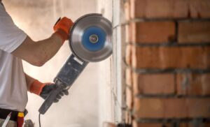 A man wearing a plain white shirt and black and orange gloves while using an angle grinder to cut fire bricks on a wall