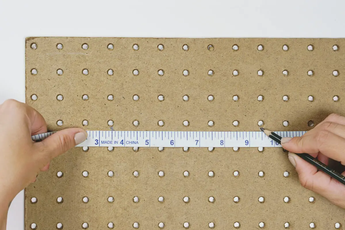 A person using a white measuring tape and black pencil to measure a brown peg board