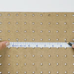 A person using a white measuring tape and black pencil to measure a brown peg board