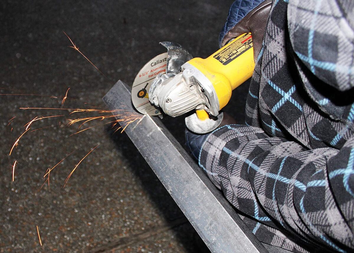 A person wearing a black, blue, and gray checkered long sleeves shirt is using a yellow angle grinder to cut silver steel near the brown tiles