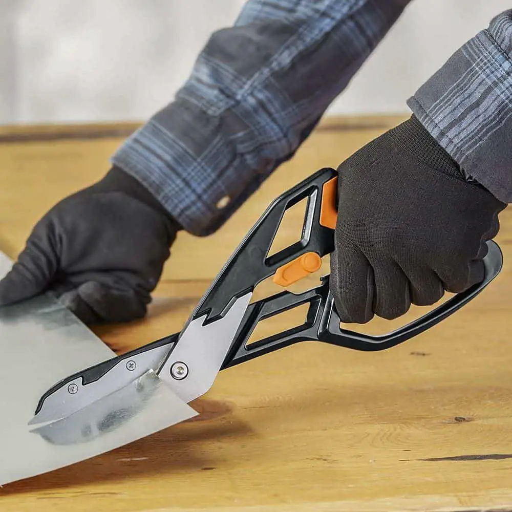 A person wearing a checkered long sleeves shirt and black gloves is using a black metal cutting snips to cut a sheet of metal on top of a brown wooden table