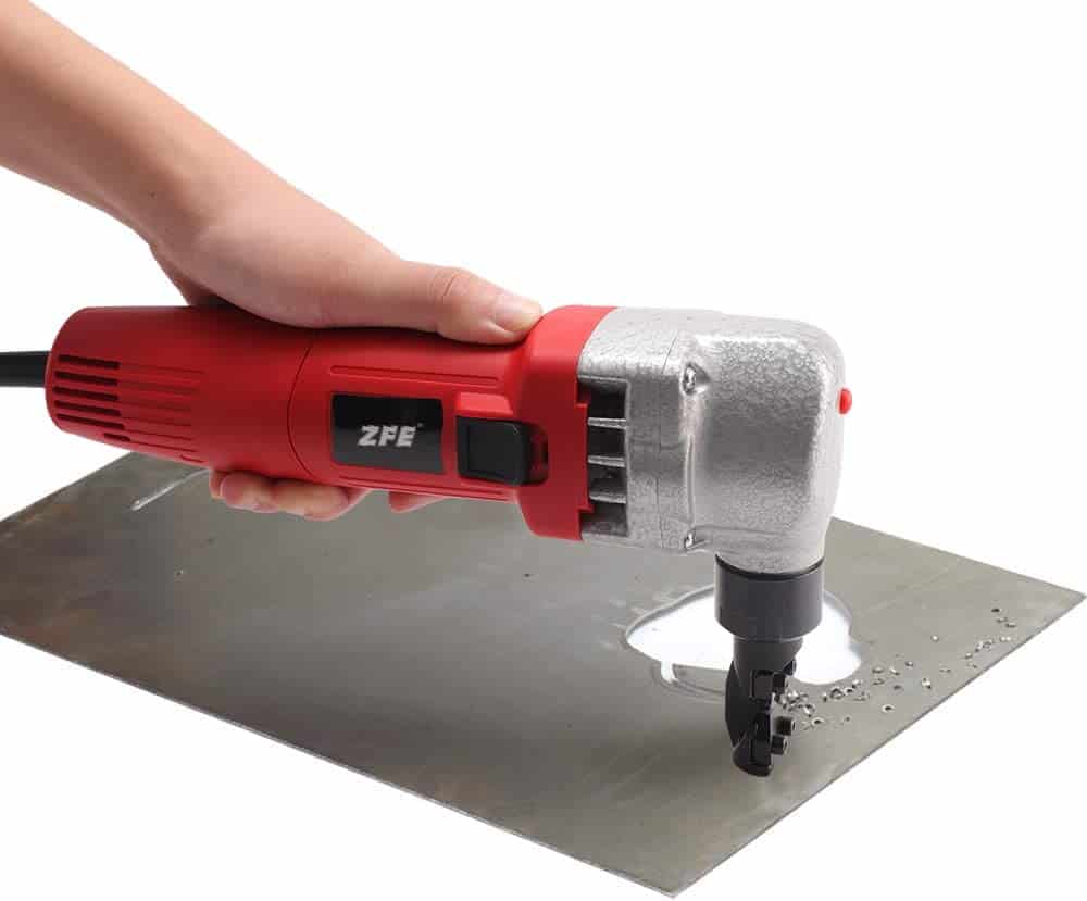 A person using a red nibbler to cut a silver metal sheet on top of a white surface