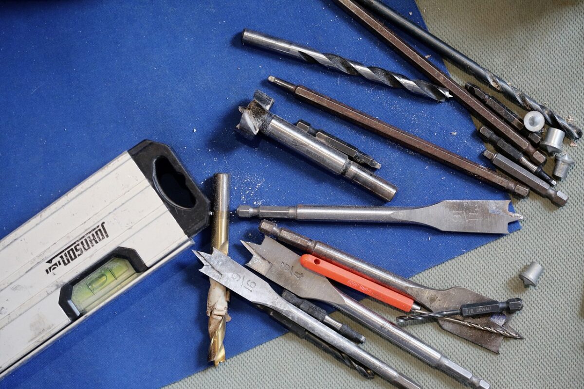 Different sizes of drills near a silver and black colored level on top of blue sandpaper on a gray table