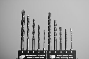Different sizes of wood drills on a black container near a gray wall