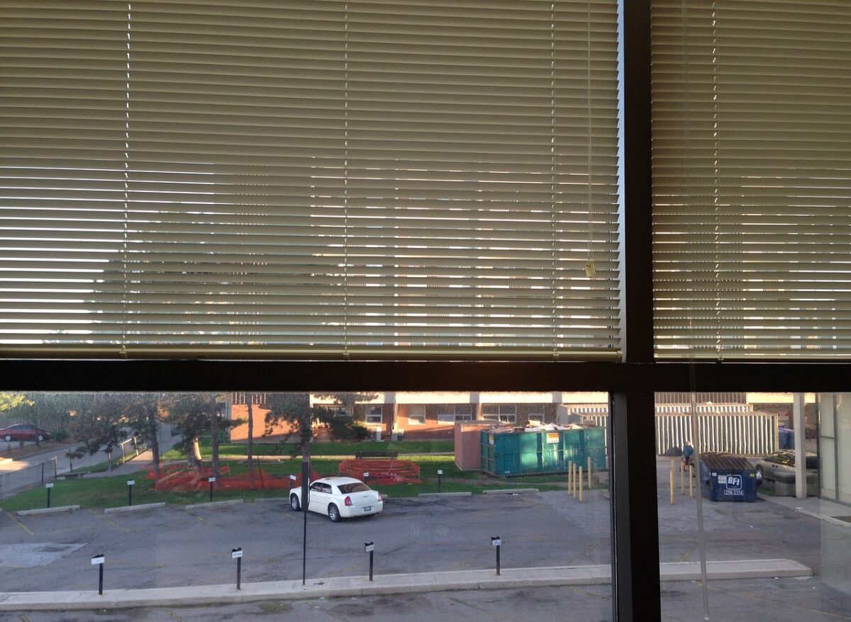 A window with the upper part having blinds while the lower part shows the view outside with a parking lot and a white car