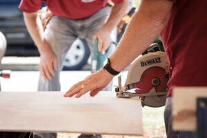 A man cutting a thin plywood using a circular saw with a man on background holding another power tool