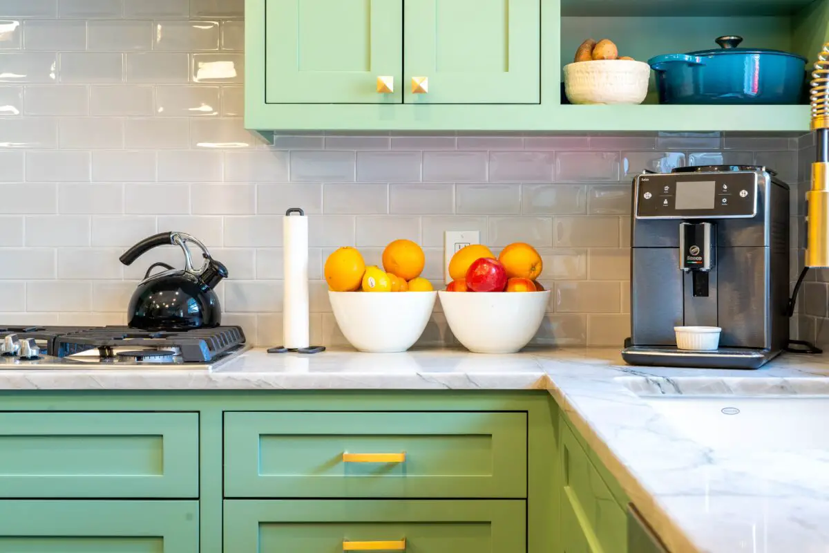 A countertop with marble design and green cabinets together with a coffee maker, two bowl of fruits, paper towel, and a stovetop with kettle